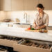 Cuisine contemporaine lumineuse avec grand plan de travail en pierre et rangement soigné, une femme prépare des ingrédients sur une planche à découper.
