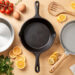 Vue du dessus d’une cuisine lumineuse présentant trois poêles, en inox, en fonte et antiadhésive, entourées d’ingrédients frais et d’une spatule en bois.